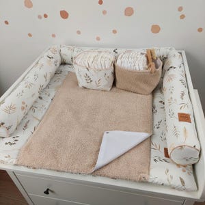 Peut inclure: Une table à langer blanche avec un matelas à langer beige et deux paniers remplis de couches. La table a un tour de lit à imprimé floral et une brosse en bois. Le mur derrière la table a des pois marron et orange.