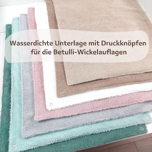 Könnte beinhalten: Stapel wasserdichter Wickelauflagen in verschiedenen Pastellfarben, darunter Grün, Blau, Rosa, Grau, Weiß und Beige. Jedes Pad hat Druckknöpfe. Der Text auf dem Bild lautet: "Wasserdichte Unterlage mit Druckknöpfen für die Betulli-Wickelauflagen."