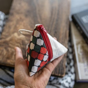 Könnte beinhalten: Kleine, dreieckige Tasche mit rotem Reißverschluss und weißer Schlaufe. Die Tasche hat ein geometrisches Muster aus roten, grünen und schwarzen Formen auf weißem Grund. Die Tasche wird in einer Hand gehalten.