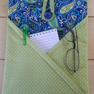 May include: A green fabric pouch with white polka dots, featuring a flap with a blue paisley pattern. The pouch has a blue button closure and contains a notepad, a pen, and a pair of glasses.