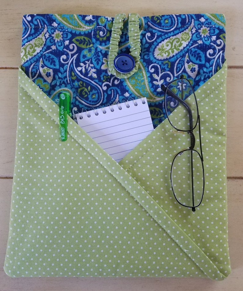 May include: A green fabric pouch with white polka dots, featuring a flap with a blue paisley pattern. The pouch has a blue button closure and contains a notepad, a pen, and a pair of glasses.