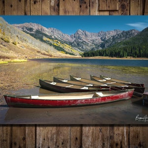 May include: Scenic landscape photograph featuring a lake surrounded by mountains and trees under a clear blue sky. Several canoes are pulled up on the shore, with a red canoe in the foreground. The image is framed by a wooden border.
