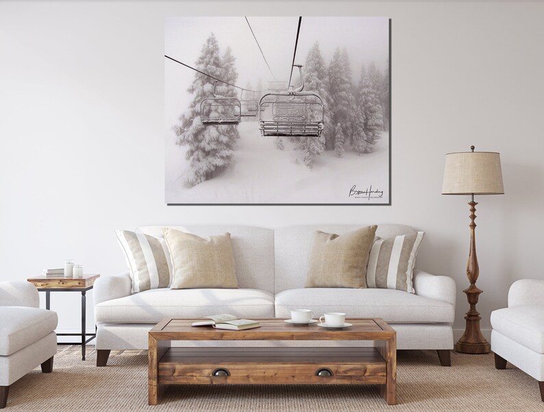 May include: Black and white photograph of a ski lift ascending through a snowy forest. The image features a row of ski lift chairs, with snow-covered evergreen trees in the background. The artist's signature is visible in the lower right corner.