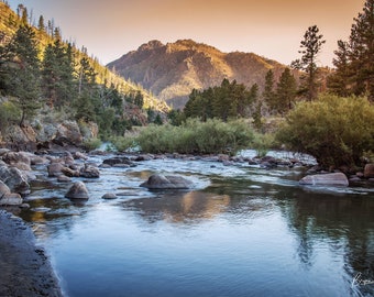 May include: A scenic view of a river flowing through a rocky landscape. The river is calm and clear, with a few rocks visible in the water. The surrounding landscape is covered in trees and mountains, with a golden sunset in the background.