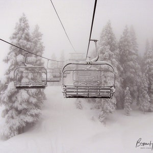 May include: A black and white photograph of a ski lift with empty chairs suspended by cables. The lift ascends through a snow-covered forest, with frosted evergreen trees and a foggy atmosphere. The image captures a serene winter scene.