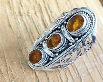 May include: A silver ring with an intricate design and three amber gemstones. The ring is set on a wooden surface.