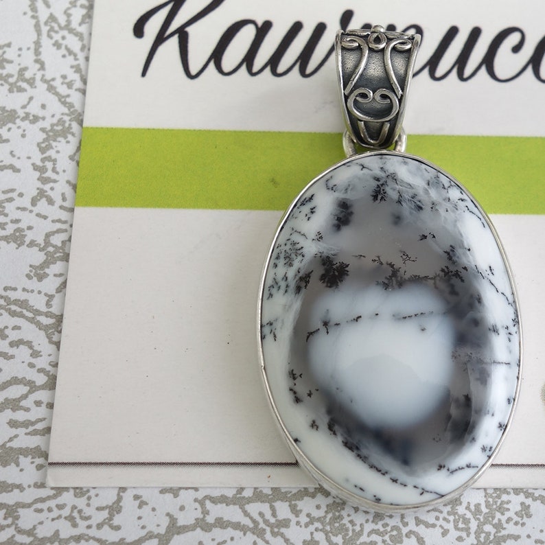 May include: A silver pendant with an oval-shaped dendritic agate stone. The stone is white with black dendritic patterns. The pendant has a silver bail.