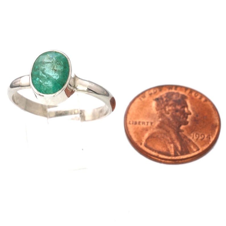 May include: A silver ring with a round, green emerald gemstone. The ring is shown next to a US penny for size comparison.