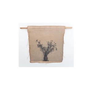 Puede incluir: Un tapiz de tela beige con una ilustración en blanco y negro de un jarrón con flores secas. La tela está sujeta a una varilla de madera. Los bordes de la tela están deshilachados.