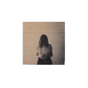 Puede incluir: Una impresión artística cuadrada que presenta una fotografía en tono sepia de una persona vista desde atrás. La persona tiene el cabello largo y sostiene un objeto grande y claro en forma de pluma o abanico contra su torso, sobre un fondo texturizado.