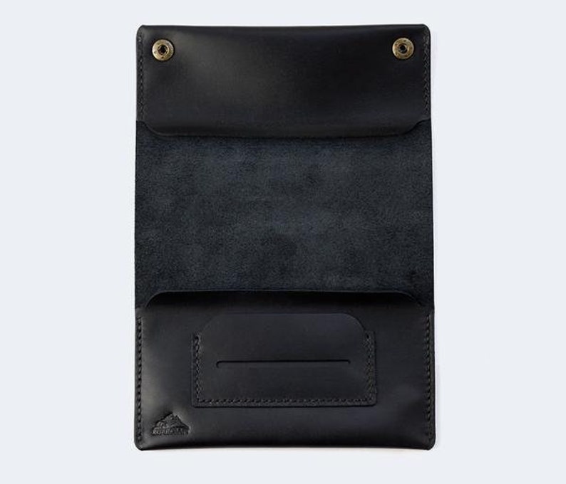 Puede incluir: Una cartera de cuero negro con cierre de solapa y un peque&ntilde;o bolsillo en la parte delantera. La cartera tiene un logotipo en relieve sutil en la parte delantera.