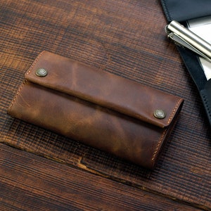 Puede incluir: Una cartera de cuero marr&oacute;n con una solapa plegable y dos broches met&aacute;licos. La cartera est&aacute; sobre una superficie de madera.