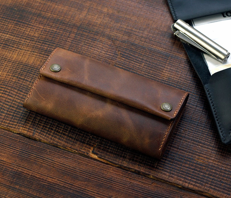 Puede incluir: Una cartera de cuero marr&oacute;n con una solapa plegable y dos broches met&aacute;licos. La cartera est&aacute; sobre una superficie de madera.