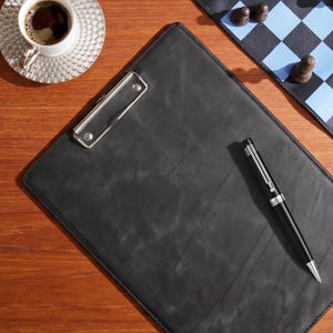 Könnte beinhalten: Ein schwarzes Klemmbrett aus Leder mit einer silbernen Metallklemme an der Oberseite, begleitet von einem eleganten schwarzen und silbernen Kugelschreiber, der auf der Oberfläche liegt. Das Klemmbrett hat eine glatte, matte Textur und ist rechteckig.