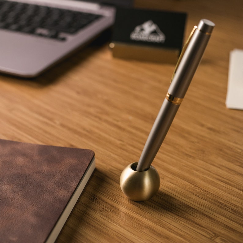 May include: A gold pen holder with a silver pen resting inside. The pen holder is a sphere and the pen is a classic design with a gold band.
