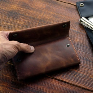 Puede incluir: Una cartera de cuero marr&oacute;n con cierre de solapa y m&uacute;ltiples cierres a presi&oacute;n. La cartera est&aacute; abierta y el interior es visible.