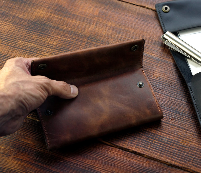 Puede incluir: Una cartera de cuero marr&oacute;n con cierre de solapa y m&uacute;ltiples cierres a presi&oacute;n. La cartera est&aacute; abierta y el interior es visible.