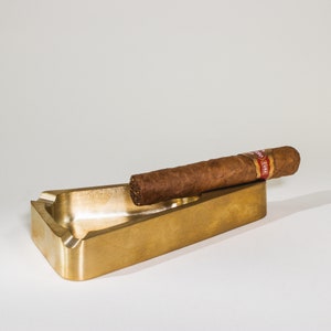 May include: A gold-colored metal cigar ashtray with a cigar resting on the edge. The cigar has a red and white band that says "CUBA".