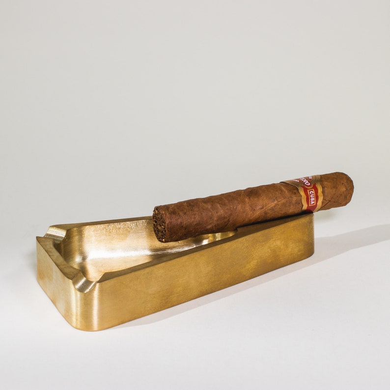 May include: A gold-colored metal cigar ashtray with a cigar resting on the edge. The cigar has a red and white band that says "CUBA".