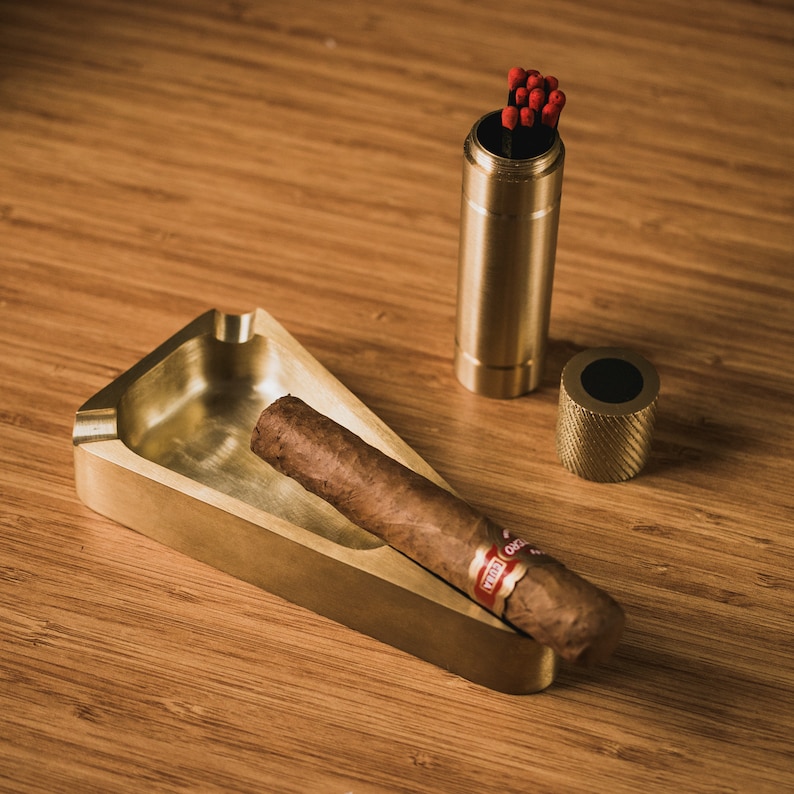 May include: A brass cigar ashtray with a triangular shape and a cigar resting inside. A brass match holder with a black top and a stack of red matches is also visible.