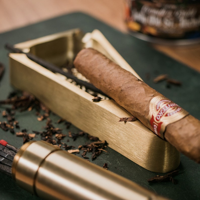 May include: A gold-colored metal cigar ashtray with a brown cigar resting on top. The cigar has a red and white band with the text "Montecristo" printed on it.