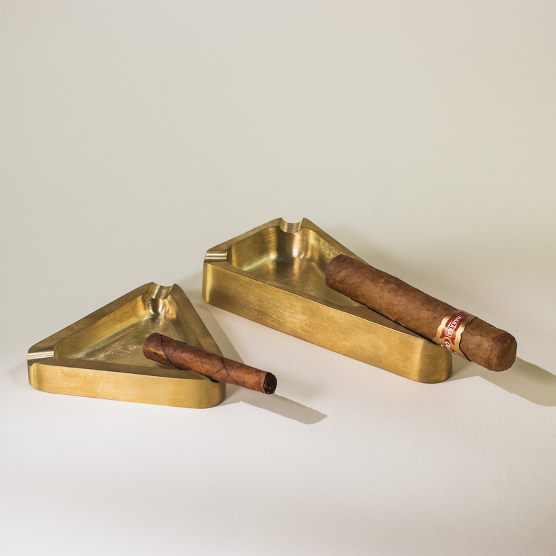 May include: Two gold-colored triangular ashtrays with a cigar resting in each. The ashtrays are made of metal and have a smooth, polished finish.