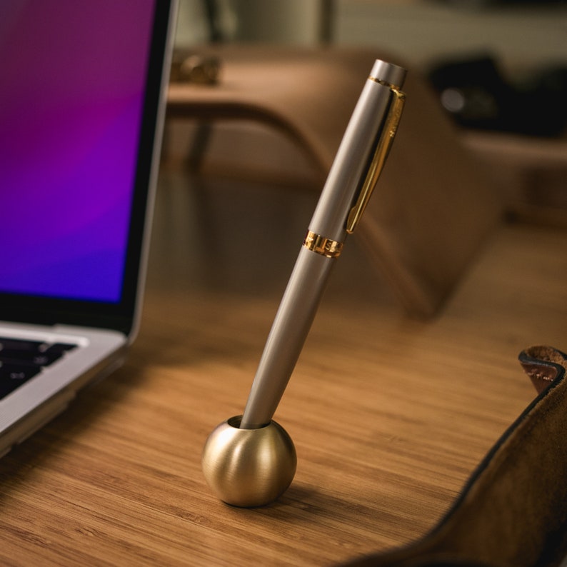 May include: A silver pen with a gold accent is placed in a round, silver pen holder. The pen holder is on a wooden surface.