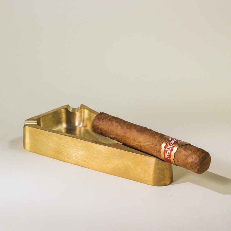 May include: A gold-colored metal ashtray with a cigar resting inside. The cigar has a red and white band with the word "CUBA" printed on it.