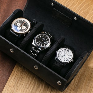 May include: A black leather watch roll with four slots for storing watches. The roll is open and three watches are visible: a silver chronograph watch, a silver watch with a black face, and a silver watch with a white face.