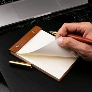 Pode incluir: Um bloco de notas em couro castanho com papel pontilhado. O bloco de notas está aberto, com uma página a ser virada. Uma caneta de madeira e uma caneta dourada também são visíveis. O bloco de notas está numa superfície preta.
