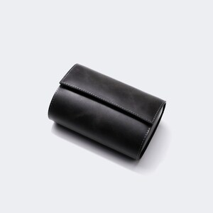 May include: A black leather watch roll with a stitched seam. The roll is closed and lying on a white surface.