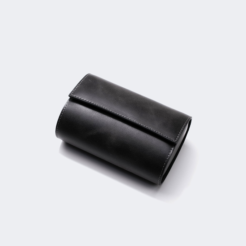 May include: A black leather watch roll with a stitched seam. The roll is closed and lying on a white surface.