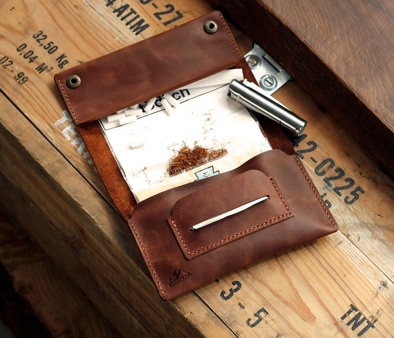Puede incluir: Una bolsa de tabaco de cuero marr&oacute;n con una solapa que se abre para revelar un compartimento para papeles de liar, un compartimento para tabaco y un peque&ntilde;o bolsillo para un encendedor. La bolsa tiene un cierre de presi&oacute;n met&aacute;lico y un peque&ntilde;o logotipo met&aacute;lico en la parte delantera.