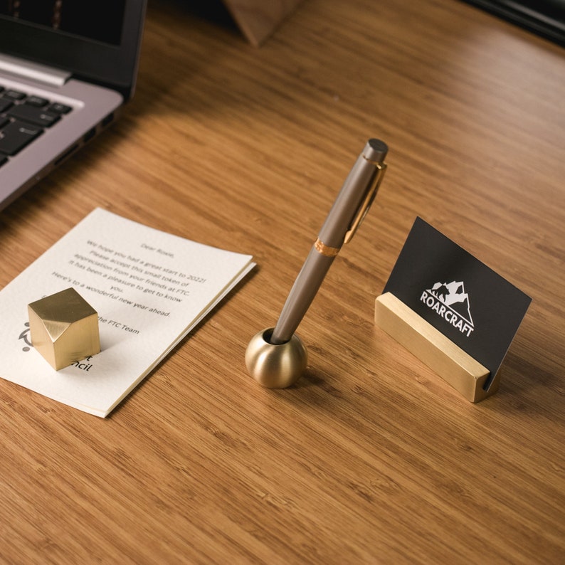 May include: A gold metal pen holder with a round top and a silver pen. A black business card holder with the text "ROARCRAFT" is on a wooden surface.