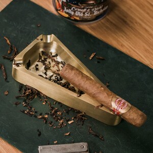 May include: A gold-colored ashtray with a triangular shape and a cigar resting inside. The ashtray is filled with tobacco shavings. The cigar is labeled "Montecristo"