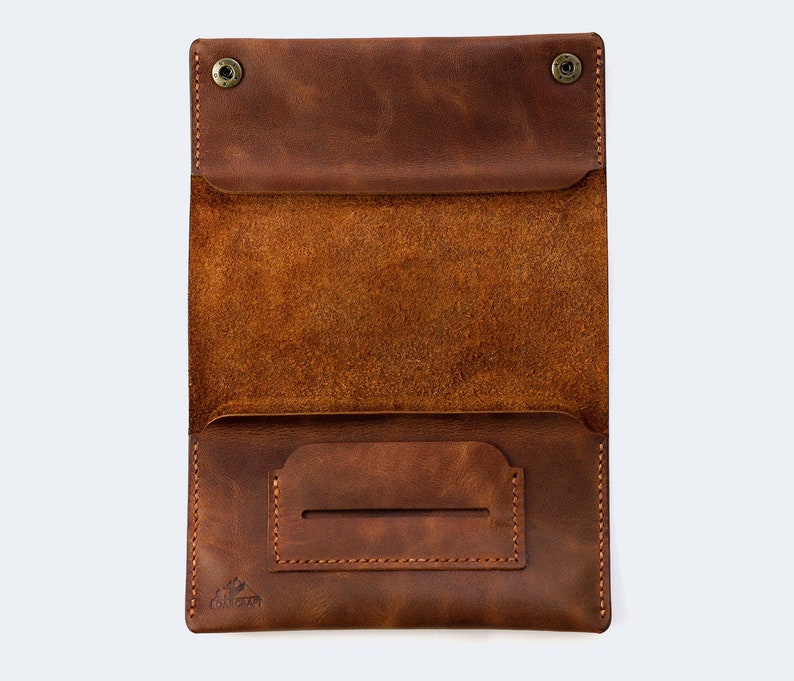 Puede incluir: Una cartera de cuero marr&oacute;n con una solapa y un peque&ntilde;o bolsillo en la parte delantera. La cartera est&aacute; abierta y el interior es visible. El interior de la cartera tiene un bolsillo grande y un bolsillo m&aacute;s peque&ntilde;o en la parte delantera. La cartera tiene un peque&ntilde;o logotipo en la parte delantera que dice "COUCHCRAFT".