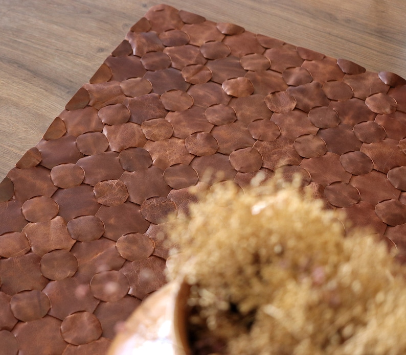 May include: A brown leather rug with a woven pattern of circles. The rug is made of individual pieces of leather that are sewn together.
