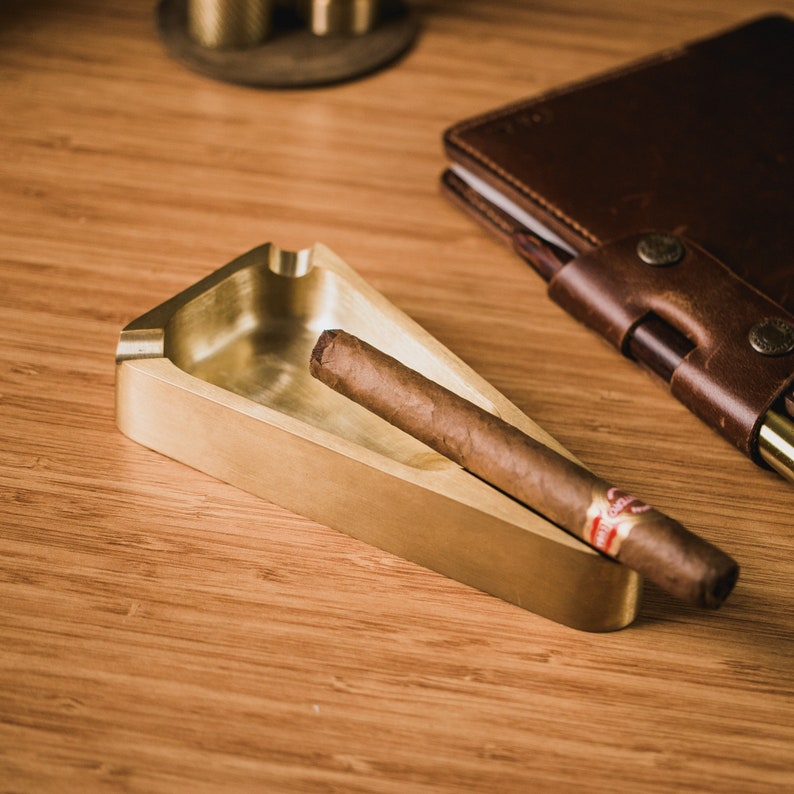 May include: A gold-colored triangular ashtray with a cigar resting inside. The ashtray is on a wooden surface.