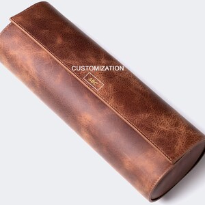May include: A brown leather cylindrical roll-up case with a flap closure. The flap has a gold embossed inscription that reads "CUSTOMIZATION ABC".