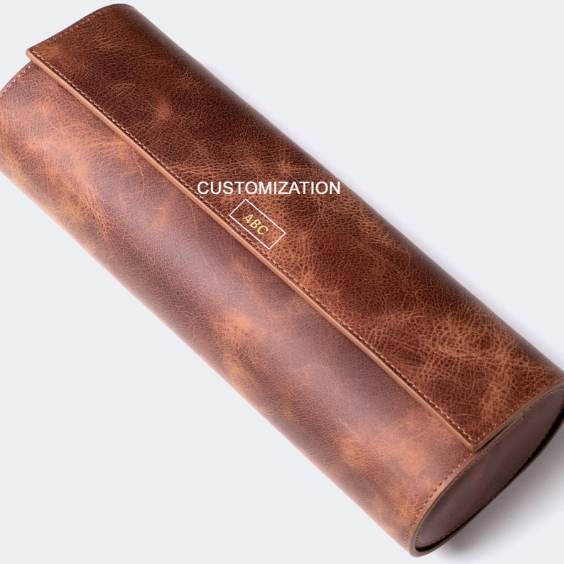 May include: A brown leather cylindrical roll-up case with a flap closure. The flap has a gold embossed inscription that reads "CUSTOMIZATION ABC".