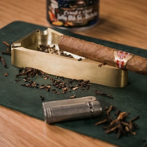 May include: A brass cigar ashtray with a dark green felt base. A cigar rests in the ashtray, and a silver lighter sits on the felt base. There are tobacco shavings scattered around the ashtray.