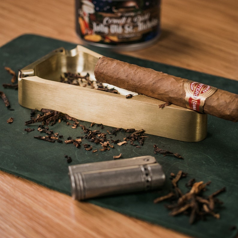May include: A brass cigar ashtray with a dark green felt base. A cigar rests in the ashtray, and a silver lighter sits on the felt base. There are tobacco shavings scattered around the ashtray.