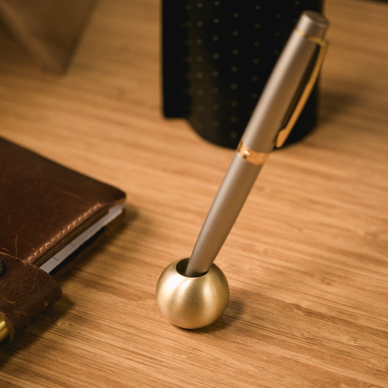 May include: A silver pen with a gold band sits in a gold metal pen holder. The pen holder is round and has a smooth surface.