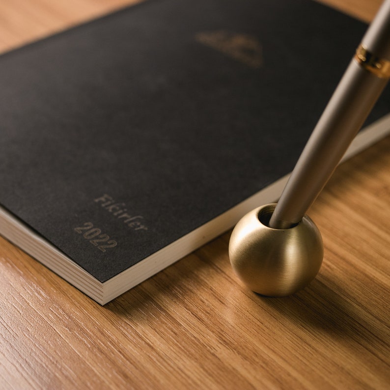 May include: A black notebook with the text "Ficirler 2022" on the cover, a gold pen holder, and a silver pen.