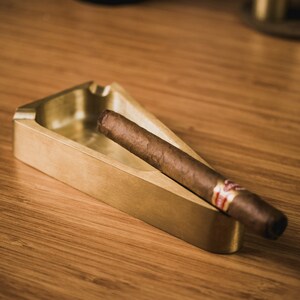 May include: A gold-colored metal ashtray with a triangular shape and a cigar resting inside. The ashtray is on a wooden surface.