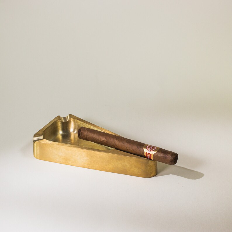 May include: A gold-colored metal ashtray with a cigar resting inside. The ashtray is shaped like a triangle with rounded edges.
