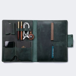 May include: A dark green leather portfolio with multiple pockets and compartments. The portfolio is open and contains a phone, pens, a notebook, and a charging cable.