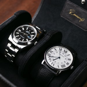 May include: Two luxury wristwatches in a black watch box. The watch on the left has a silver band and a black face with silver hands. The watch on the right has a black leather band and a white face with black Roman numeral markings and silver hands. The text "CARTIER" is visible on the watch face.