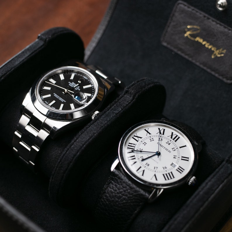 May include: Two luxury wristwatches in a black watch box. The watch on the left has a silver band and a black face with silver hands. The watch on the right has a black leather band and a white face with black Roman numeral markings and silver hands. The text "CARTIER" is visible on the watch face.
