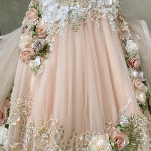Puede incluir: Un vestido de tul rosa claro con intrincados bordados florales y aplicaciones florales 3D. El vestido presenta una variedad de flores en tonos de blanco, rosa y verde.